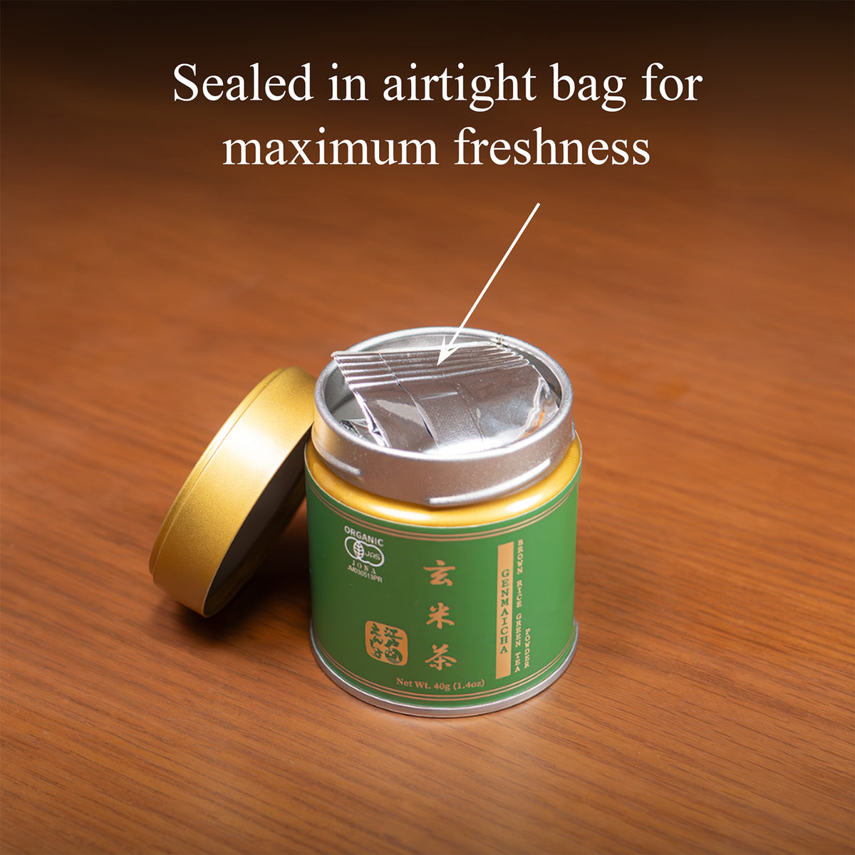 Open Genmaicha tin on a wooden surface with the gold lid beside it, revealing the inner foil bag, text 'Sealed in airtight bag for maximum freshness'.
