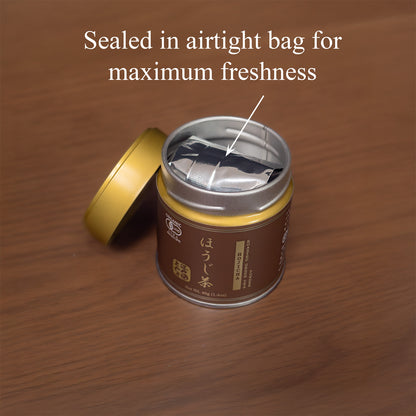 Open Hojicha tin on a wooden surface with the gold lid beside it, revealing the inner foil bag, text 'Sealed in airtight bag for maximum freshness'.