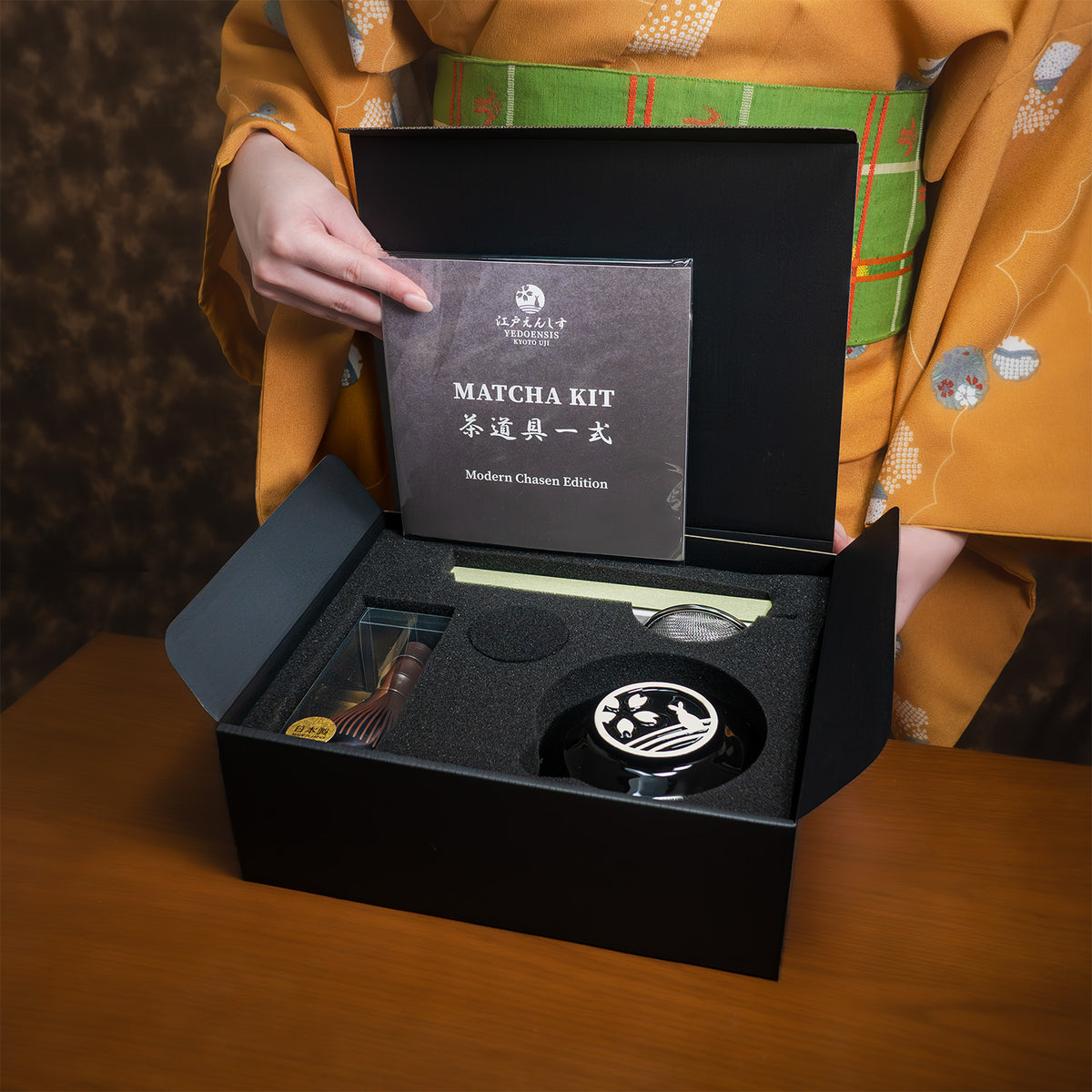 A woman in an orange kimono opening the black Matcha Kit box on a wooden table, lifting the 'Matcha Kit - Modern Chasen Edition' insert card above the foam tray holding the bowl, whisk and sifter.