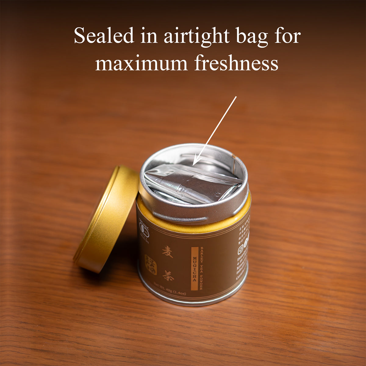 Open Mugicha barley tea tin on a wooden surface with the gold lid beside it, revealing the inner foil bag, text 'Sealed in airtight bag for maximum freshness'.