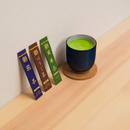 Sencha, Hojicha and Genmaicha sticks leaning against a wall beside a navy cup of green tea on a cork coaster.