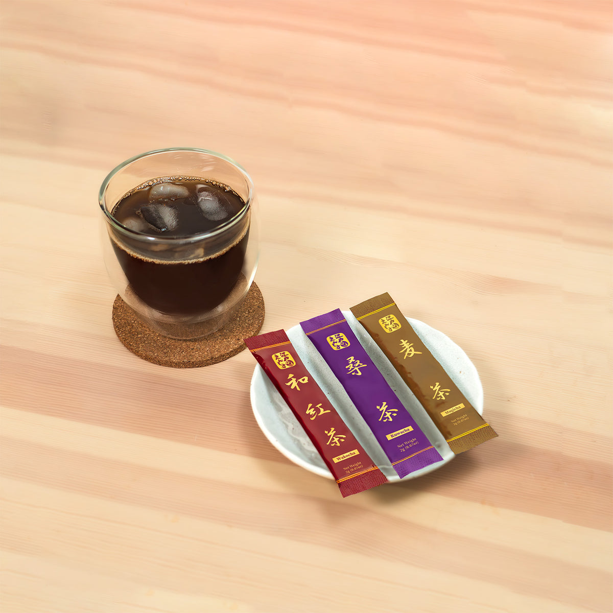 Wakocha, Kuwacha and Mugicha sticks on a white dish beside a glass of iced brown tea on a cork coaster.