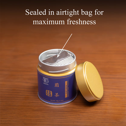 Open Sencha tin on a wooden surface with the gold lid beside it, revealing the inner foil bag, text 'Sealed in airtight bag for maximum freshness'.